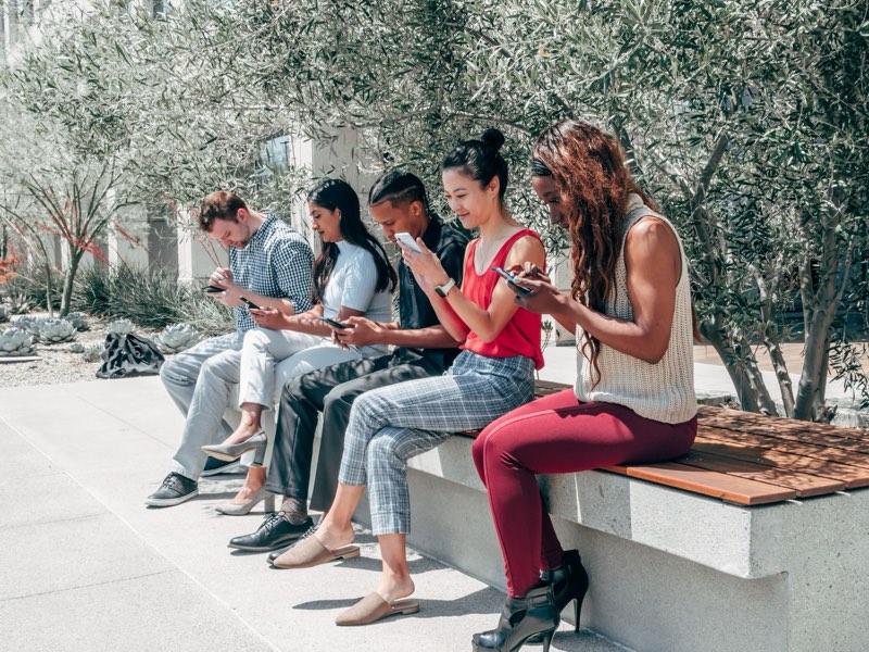 Pessoas sentadas num banco de praça, todas usando o celular